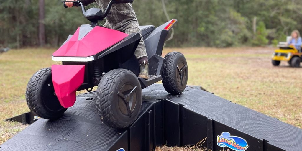 Child riding a toy vehicle over a small ramp outdoors.